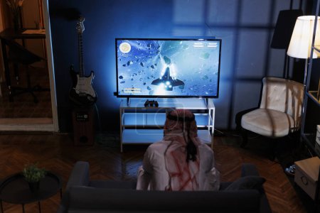 Téléchargez les photos : Homme du Moyen-Orient en tenue traditionnelle jouant un jeu vidéo immersif dans le confort de sa maison. L'écran détaillé et le contrôleur sans fil améliorent son expérience de jeu. - en image libre de droit