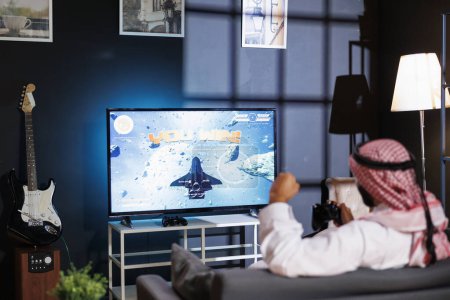Photo for Middle Eastern man in traditional attire celebrates a victory, immersed in virtual reality game. With wireless controller, he grasps the joystick, capturing the excitement of online gaming. - Royalty Free Image
