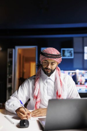 Foto de Chico musulmán profesional utiliza la tecnología para trabajar eficientemente en la oficina, navegar en línea y escribir notas desde un ordenador portátil. Estudiante árabe joven usando su cuaderno y minicomputador para la investigación. - Imagen libre de derechos