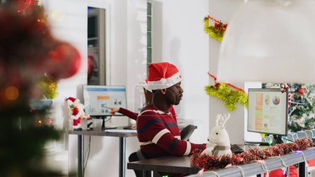 Foto de Empleado afroamericano con sombrero de Santa Claus trabajando en la oficina adornada de Navidad, sintiéndose cansado. El miembro del personal que bebe café mientras revisa los números de ingresos empresariales en el lugar de trabajo de Navidad adornan - Imagen libre de derechos