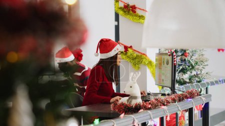 Photo for Revealing shot of focused asian employee working on christmas season in decorated office. Diligent worker solving tasks at computer desk in diverse xmas ornate workplace next to BIPOC colleagues - Royalty Free Image