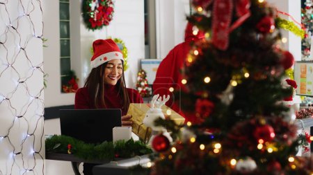 Photo for Asian worker receiving gifts from generous colleague acting as Santa in xmas decorated office. Employee wearing festive costume surprising coworkers with presents during winter holiday season - Royalty Free Image