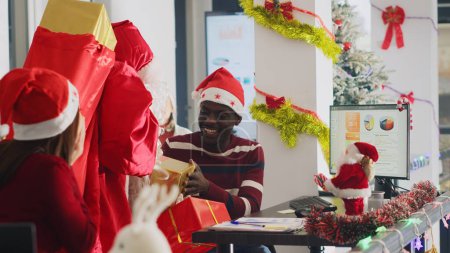 Téléchargez les photos : Heureux travailleur afro-américain recevant des cadeaux du Père Noël dans l'espace de travail orné de Noël. Employé excité obtenir des cadeaux de collègue déguisé avant Noël fête de bureau - en image libre de droit