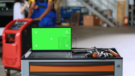 Photo for Green screen laptop placed on working bench in busy garage next to professional tools while mechanics walk around in blurry background. Chroma key device in auto repair shop, close up - Royalty Free Image