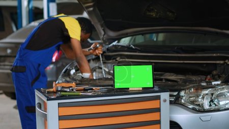 Photo for Engineer in car service uses torque wrench to secure bolts inside vehicle with green screen tablet in front. Expert utilizes professional tools to mend out of order automobile next to mockup device - Royalty Free Image