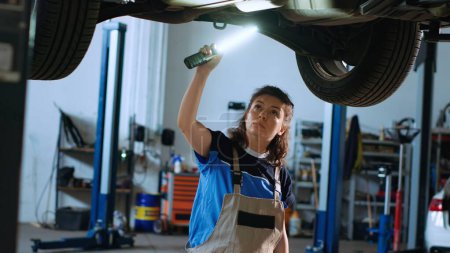 Photo for Mechanic underneath vehicle on overhead lift in garage, checking parts during annual checkup. Car service professional using work light to make sure automotive underbody is in proper condition - Royalty Free Image