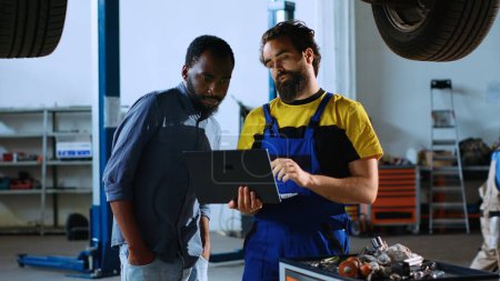 Photo for Licensed repairman in garage workspace standing with customer underneath suspended car, looking together for replacement parts. Worker helps man fix vehicle, using laptop to find components - Royalty Free Image