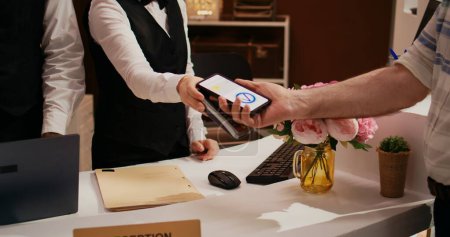 Photo for Elderly man makes nfc transaction with mobile phone at pos terminal, paying for accommodation at five star resort. Tourist arriving at front desk doing check in for holiday. Close up. Handheld shot. - Royalty Free Image