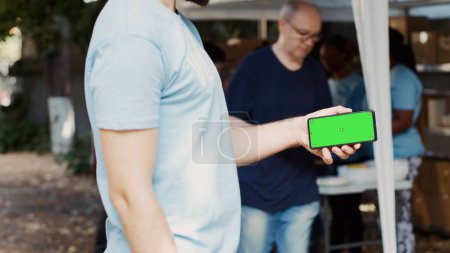 Photo for Detailed view of person horizontally holding mobile phone at charitable food drive, presenting an isolated mockup template. Volunteer grasping smartphone with blank green screen display. Tripod shot. - Royalty Free Image