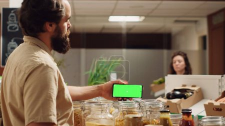 Foto de Hombre usando el teléfono inteligente de modo de paisaje de pantalla verde en el supermercado ecológico. Cliente en la tienda de comestibles local utiliza el teléfono clave de croma mientras que las compras de verduras orgánicas, tiro panorámica - Imagen libre de derechos