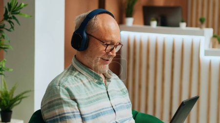 Photo for Retired traveller working on tablet in lounge area, passing time watching tv show on online streaming services. Senior man in hotel lobby using headphones and gadget, waiting to start vacation. - Royalty Free Image