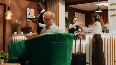 Photo for Old woman filling in registration forms at front desk, passing room check in and preparing to start retirement vacation trip. Traveller asking receptionist about all inclusive service. Handheld shot. - Royalty Free Image