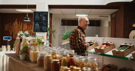 Téléchargez les photos : Un vieux client souriant à la recherche d'agrafes de garde-manger sur des étagères de supermarchés zéro déchet. Heureux homme âgé achetant des aliments biologiques dans des bocaux réutilisables à l'épicerie locale écologique - en image libre de droit