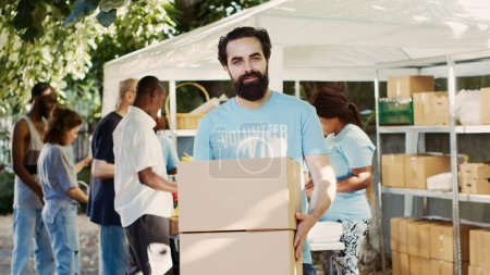 Photo for Generous man volunteering at neighborhood food drive, distributing non-perishable rations to the hungry and homeless. While holding donation box, caucasian charity worker looks at camera. Tripod shot. - Royalty Free Image