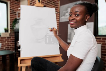 Foto de Mujer afroamericana inspirada feliz creando una obra de arte única durante el taller de arte creativo, sentada en el caballete y sonriendo a la cámara, disfrutando de la lección de dibujo, desarrollando su propia creatividad - Imagen libre de derechos