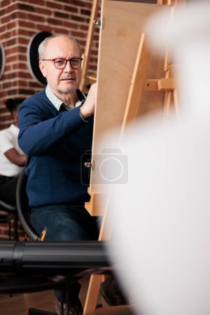 Photo for Concentrated focused senior man sitting at easel drawing object with graphite pencil. Mature student looking at ceramic vase making sketch on canvas during group art class in studio - Royalty Free Image