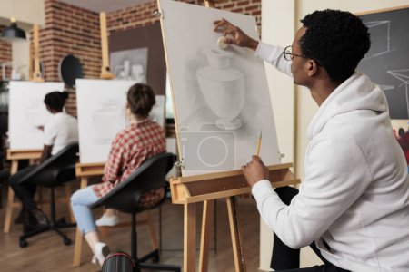 Foto de Chico afroamericano sentado en el caballete aprendiendo técnicas de dibujo, dibujando jarrón sobre lienzo mientras asistía a clases de arte en grupo creativo con amigos. Concepto de creatividad y ocio - Imagen libre de derechos