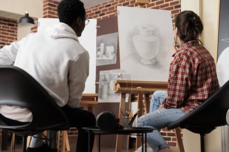 Téléchargez les photos : Jeunes étudiants diversifiés regardant toile avec vase croquis, développer des compétences en ligne et la forme tout en étudiant à l'académie d'art. Couple multiracial assis à des chevalets apprenant à se rassembler - en image libre de droit