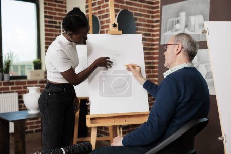 Photo for Diverse different aged art school students communicating during drawing lesson, looking at artwork on canvas and discussing sketching techniques. African American girl helping senior man at art class - Royalty Free Image