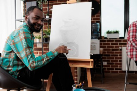 Foto de Actividades artísticas y bienestar. Retrato de chico afroamericano sonriente feliz durante la lección de dibujo, joven negro sentado en el caballete mirando a la cámara, sintiéndose inspirado durante la clase de dibujo - Imagen libre de derechos