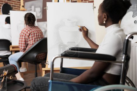 Foto de Joven mujer negra con discapacidad física visitando taller de dibujo en grupo, terapia creativa para adultos discapacitados, arte en rehabilitación física. Chica afroamericana silla de ruedas dibujo sobre lienzo - Imagen libre de derechos