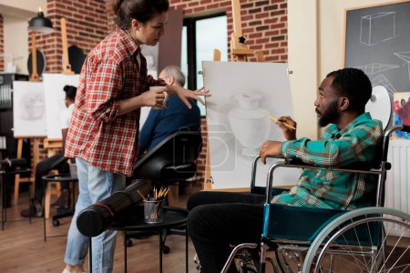 Photo for African American disabled guy talking with teacher during group drawing class, improving social skills through collaborative artwork. Teaching art to people with disabilities - Royalty Free Image