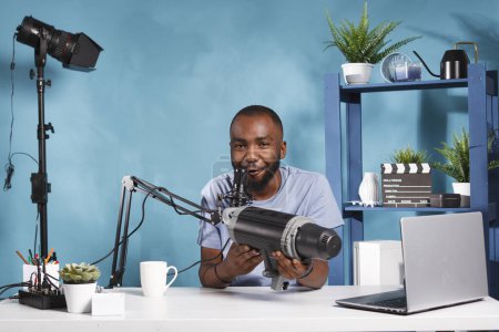 Téléchargez les photos : Vidéographe afro-américain examinant l'équipement de foudre professionnel studio sur le blog en ligne de la technologie. Jeune vlogger tenant matériel de photographie et enregistrement vidéo de présentation du produit - en image libre de droit