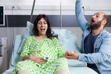 Téléchargez les photos : Jeune couple attend bébé dans le lit de service de l'hôpital à l'établissement de santé. Femme enceinte ayant des contractions douloureuses entrant dans le travail pendant que le mari panique à propos de l'accouchement. - en image libre de droit