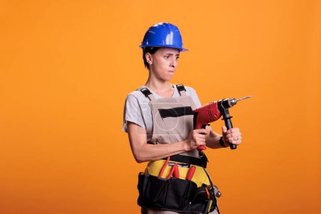 Photo for Professional female builder holding drilling gun in new interior renovation project, using power drill nail gun. Woman renovator wearing uniform in studio shot over yellow background. - Royalty Free Image