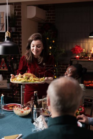 Foto de Mujer poniendo comida festiva en la mesa, disfrutando de la cena de Navidad y la celebración de temporada con diversos amigos y familiares en casa. La gente se siente feliz comiendo comida casera y bebiendo alcohol. - Imagen libre de derechos