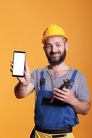 Photo for Construction worker holding cell phone with empty screen for advertising in front of camera. Professional builder with hard hat against yellow background in studio shot. - Royalty Free Image