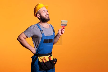 Photo for Young painter decorator painting walls with brush in studio, posing with paintbrush and tools before working on renovation. Craftsman expert using brush and color to do diy project. - Royalty Free Image