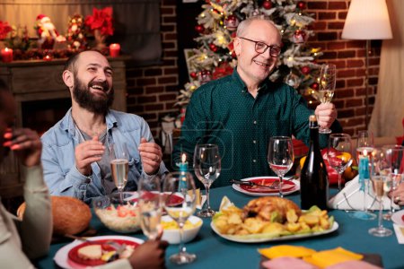 Téléchargez les photos : Un aîné porte un toast avec un verre de vin levé au dîner de Noël pour célébrer les vacances de décembre avec ses amis et sa famille. Grand-père faisant un discours autour de la table, alcool pétillant. - en image libre de droit