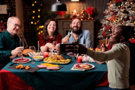 Téléchargez les photos : Les gens prenant des photos à table, faisant des souvenirs de fête de la veille de Noël réunis autour de la maison. Diverses personnes s'amusent avec du vin et de la nourriture, prenant des photos avec un appareil smartphone. - en image libre de droit