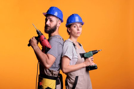 Photo for Construction workers posing with electric power drills, looking at camera and acting confident in studio. Team of builders holding drilling gun tools to work on renovation, refurbishment project. - Royalty Free Image