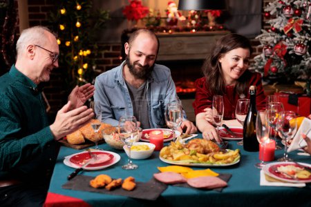 Téléchargez les photos : Les personnes âgées et les jeunes la veille de Noël se réunissant à table pour manger de la nourriture maison et boire du vin, être festif ensemble. Famille et amis divers s'amusent à célébrer les vacances de décembre. - en image libre de droit