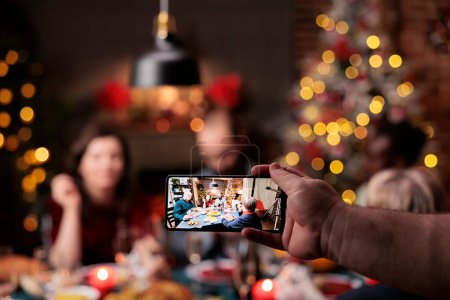 Téléchargez les photos : La personne prend des photos de personnes à table profitant de la fête du dîner avec des verres d'alcool entourés d'arbres de Noël et d'ornements. Divers amis et la famille posant pour des photos la veille de Noël. - en image libre de droit