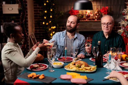 Photo for Diverse friends gathering at home to celebrate seasonal winter holiday, feeling jolly during christmas eve dinner festivity. People and family enjoying food and wine glasses, december tradition. - Royalty Free Image