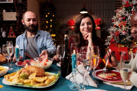 Photo for Young couple celebrating xmas with family, woman attending festive dinner during christmas eve holiday at home. Persons enjoying traditional food on the table, feeling merry and jolly. - Royalty Free Image