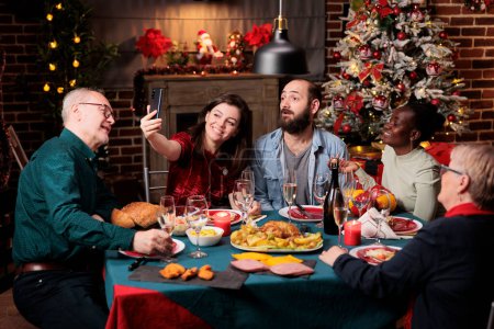 Téléchargez les photos : Les personnes appréciant les photos à la fête de la veille de Noël, célébrant les vacances de décembre ensemble autour de la table. Faire des selfies et des photos pendant le dîner, s'amuser avec un smartphone. - en image libre de droit
