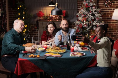 Téléchargez les photos : Des amis prenant des photos la veille de Noël, créant de beaux souvenirs de fête à la maison. Diverses personnes s'amusent avec du vin et de la nourriture, appréciant les photos pour les souvenirs à table. - en image libre de droit