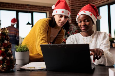 Photo for Smiling coworkers in santa hats checking annual report results on laptop in decorated festive office. African american and caucasian colleagues managing project at christmas eve - Royalty Free Image