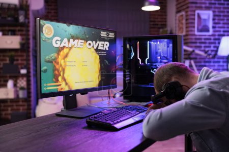 Photo for Gamer feeling gutted after losing singleplayer action videogame level, being outsmarted by enemies. Uspet man placing head on desk in frustration after seeing game over screen - Royalty Free Image