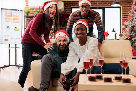 Photo for Excited employees holding gamepads and enjoying video game in festive decorated office on xmas eve. Cheerful diverse company coworkers fighting in videogame at new year holiday party - Royalty Free Image