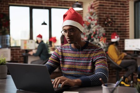 Photo for Young african american man wearing santa hat managing workload on laptop in christmas decorated office. Company employee working in workplace during winter seasonal holiday - Royalty Free Image