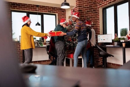 Photo for Smiling diverse colleagues exchanging christmas gifts at corporate party in decorated festive office. Company employee giving present to coworker while celebrating winter holiday at workplace - Royalty Free Image