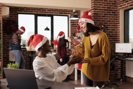 Foto de Diversas compañeras sonrientes en sombreros de Santa Claus intercambiando regalos navideños en el lugar de trabajo de la startup. Alegre empleado caucásico compartiendo bolsa de regalo festiva para compañeros de trabajo en la oficina decorada - Imagen libre de derechos