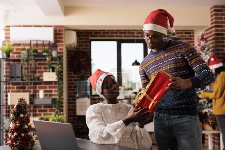Foto de Empleado de la compañía dando regalo de Navidad a su compañero de trabajo y mostrando la tarjeta de nombre adjunta a la caja. Afro-americanos colegas hombres y mujeres intercambiando regalos de Navidad en oficina decorada festiva - Imagen libre de derechos
