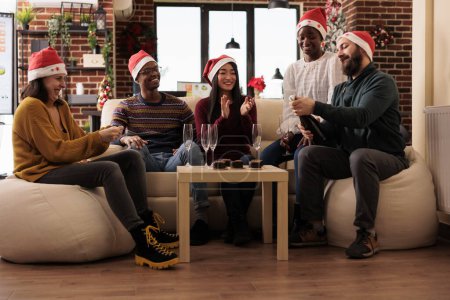 Foto de Startup trabajadores felices celebrando la Navidad, sentado alrededor de la mesa en la oficina decorada. alegre diverso equipo de compañeros de trabajo apertura botella de vino espumoso para fiesta corporativa festiva - Imagen libre de derechos