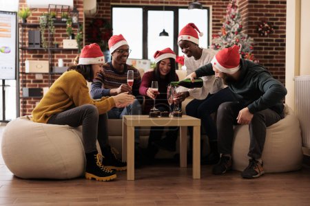 Foto de Felices compañeros de trabajo riendo y bebiendo vino espumoso mientras celebran la Navidad en un lugar de trabajo decorado. Equipo de empleados de la compañía disfrutando de vacaciones de invierno alegre celebración en la oficina - Imagen libre de derechos
